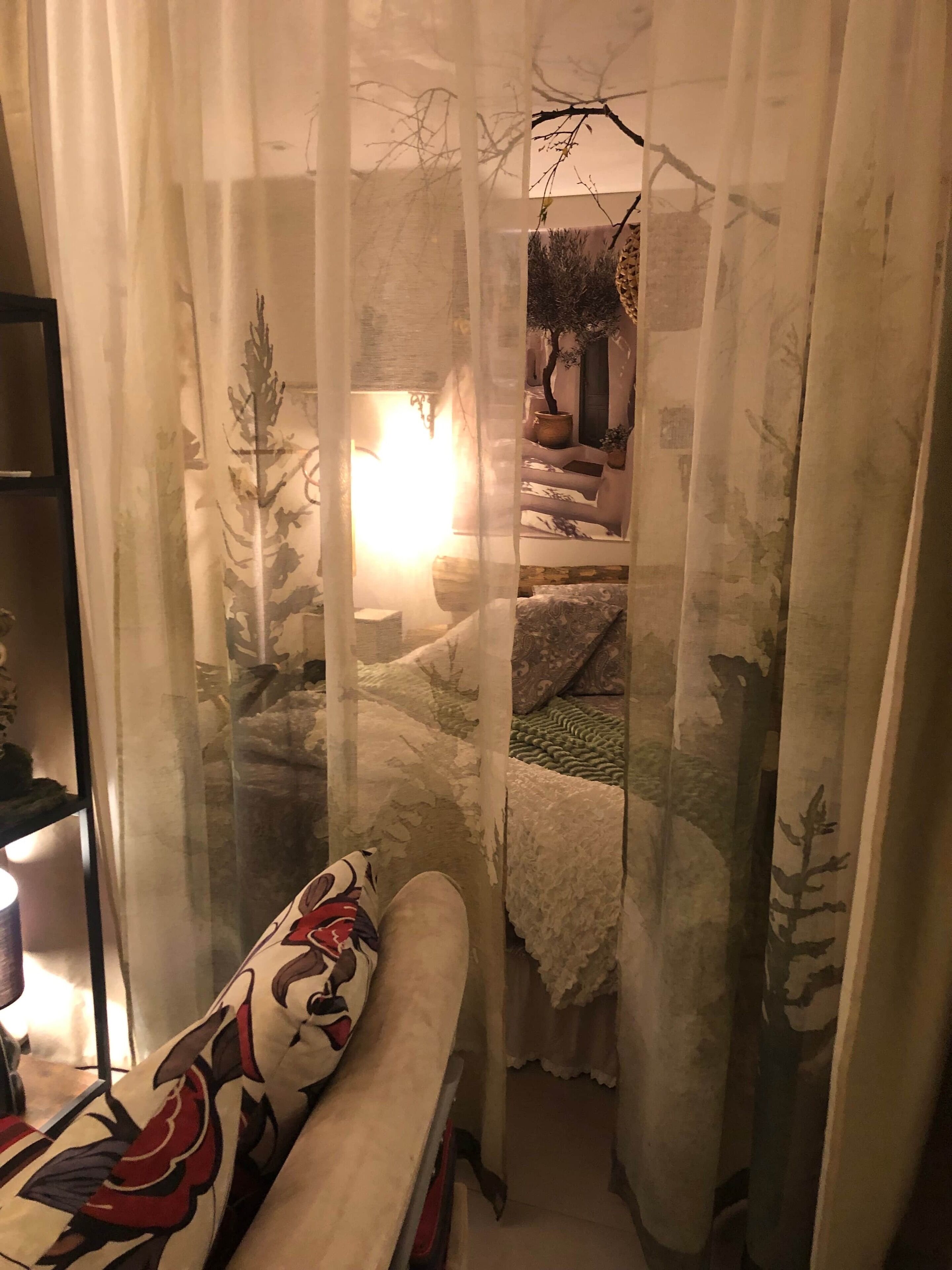 Een gezellige kamer met een bed, een stoel en een klein tafeltje, afgescheiden door dunne gordijnen met een natuurprint.