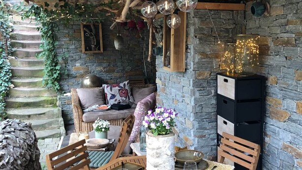 Un'area pranzo esterna rustica con un muro in pietra, mobili in legno e globi di vetro appesi.