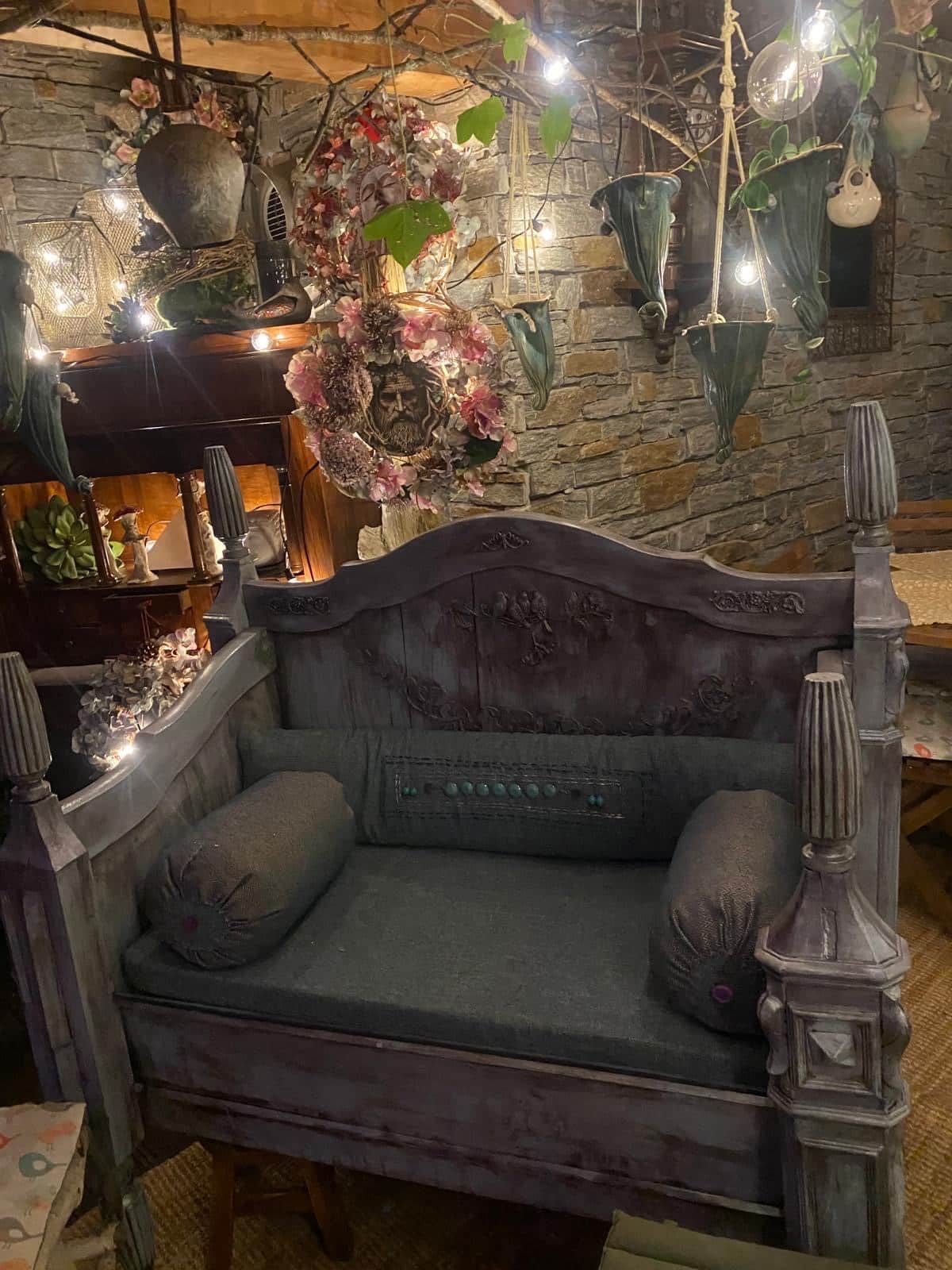 Un sofá de estilo vintage con almohadones decorativos y un cabecero de madera tallada, frente a una pared de piedra con macetas colgantes y luces de cuerda.