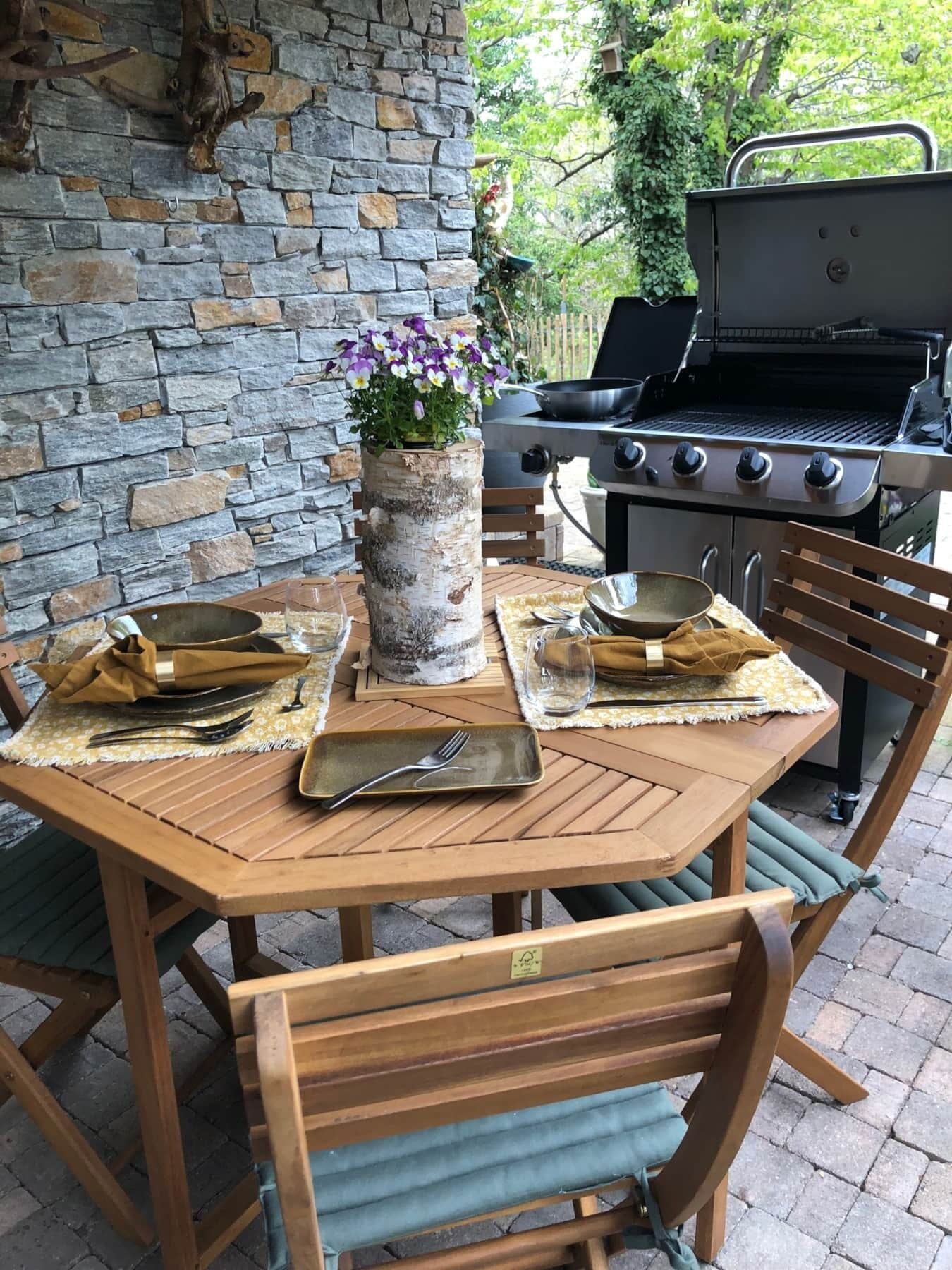 Un espace extérieur aménagé pour les repas, avec un mur en pierre, un barbecue et une table en bois dressée pour deux personnes.