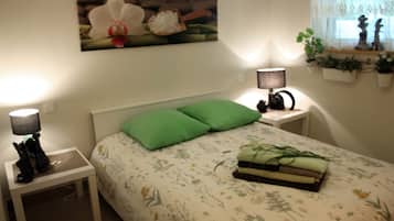 Quarto com cama, almofadas verdes, mesa de cabeceira com abajur, prateleira na parede com plantas e uma pintura de uma orquídea.