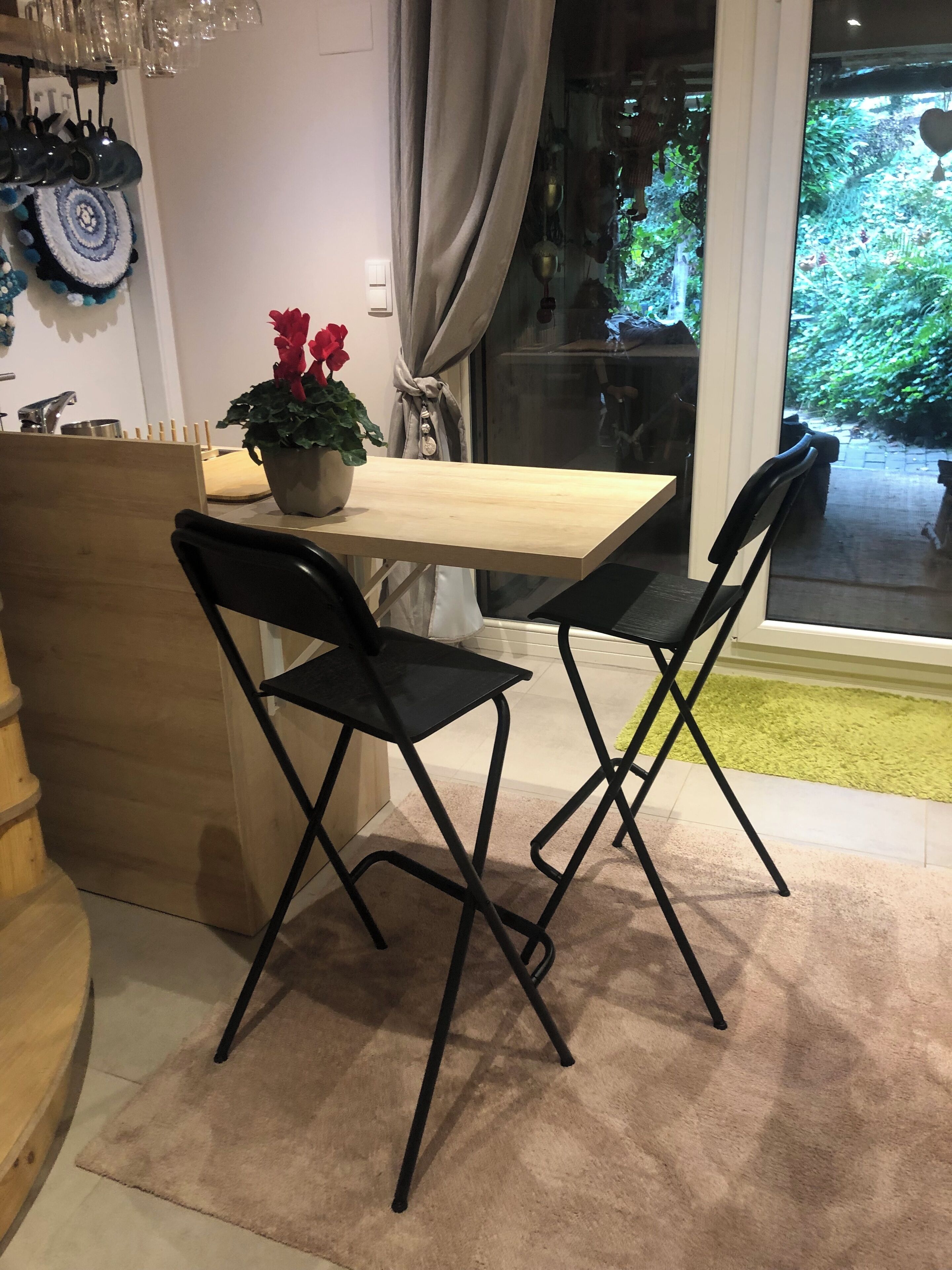 Een eetgedeelte met een houten tafel, twee zwarte stoelen, een potplant met rode bloemen en uitzicht op groen buiten het raam.