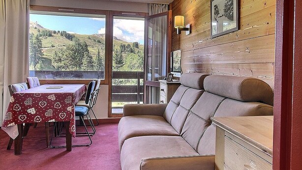 Ett rum med träinredning, en soffa, ett matbord med stolar och en utsikt över ett bergigt landskap genom stora fönster.