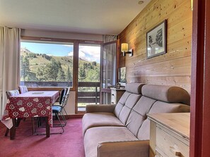 A room with a wooden interior, a sofa, a dining table with chairs, and a view of a mountainous landscape through large windows. - Ski-in/Ski-out 1-bedroom apartment, balcony, sleeps 4 (Belle Plagne)