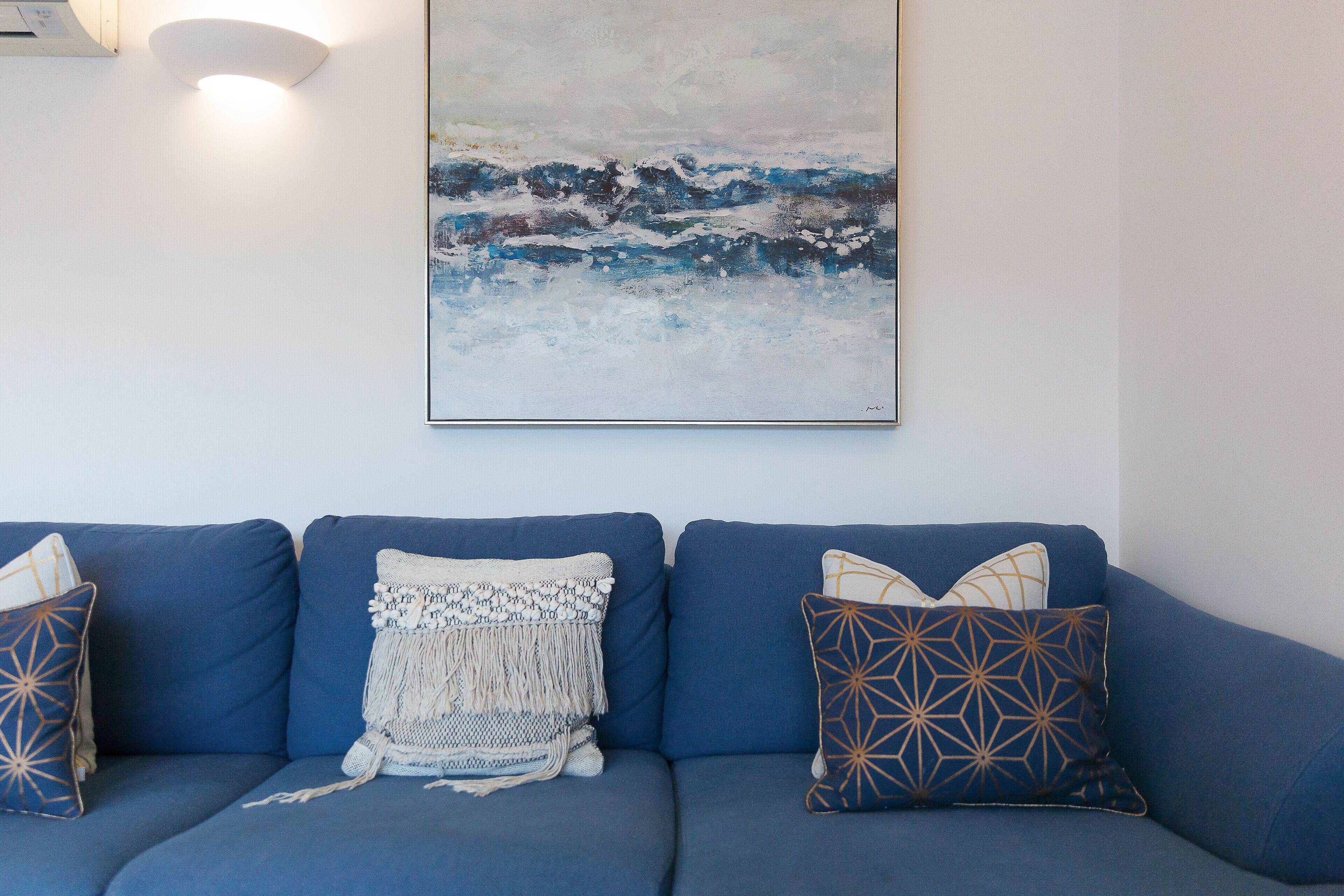 A blue sofa with patterned cushions, a wall-mounted painting, and a wall-mounted light fixture.