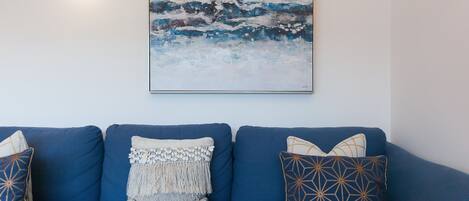 A blue sofa with patterned cushions, a wall-mounted painting, and a wall-mounted light fixture.