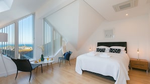 Ein modernes Schlafzimmer mit einem großen Bett, einem kleinen Tisch mit Stuhl und Blick auf die Stadt durch große Fenster.