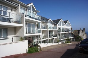 A row of modern, multi-story buildings with balconies, likely a hotel or residential complex, with a clear view of the sea in the background.