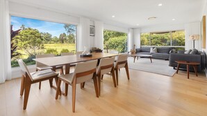 A spacious living area with a dining table and chairs, a grey sofa, and large windows offering a view of a green landscape.