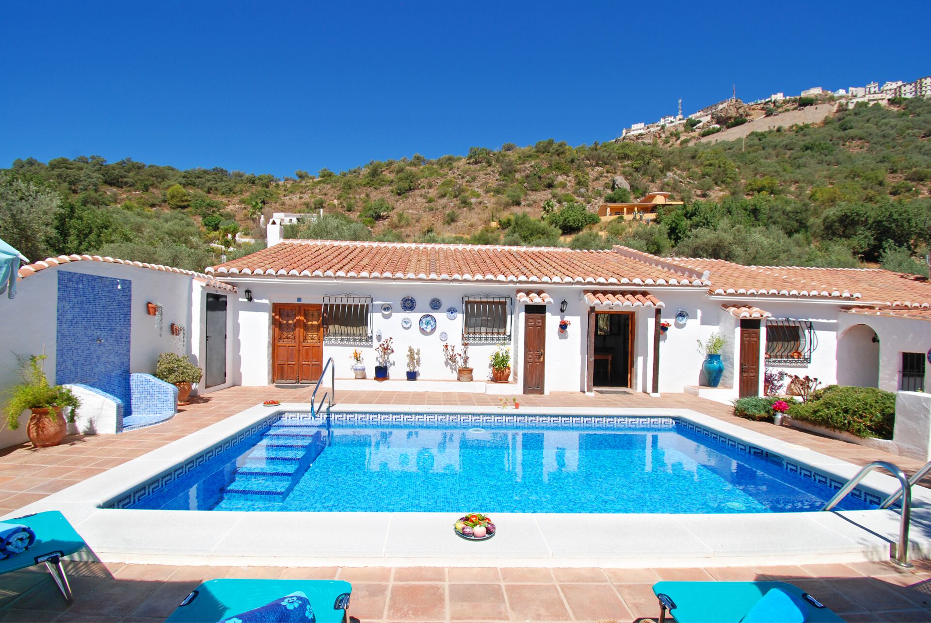 A poolside area with a swimming pool, blue lounge chairs, and a white building with a tiled roof.