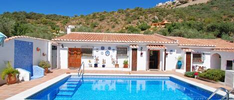 A poolside area with a swimming pool, blue lounge chairs, and a white building with a tiled roof.