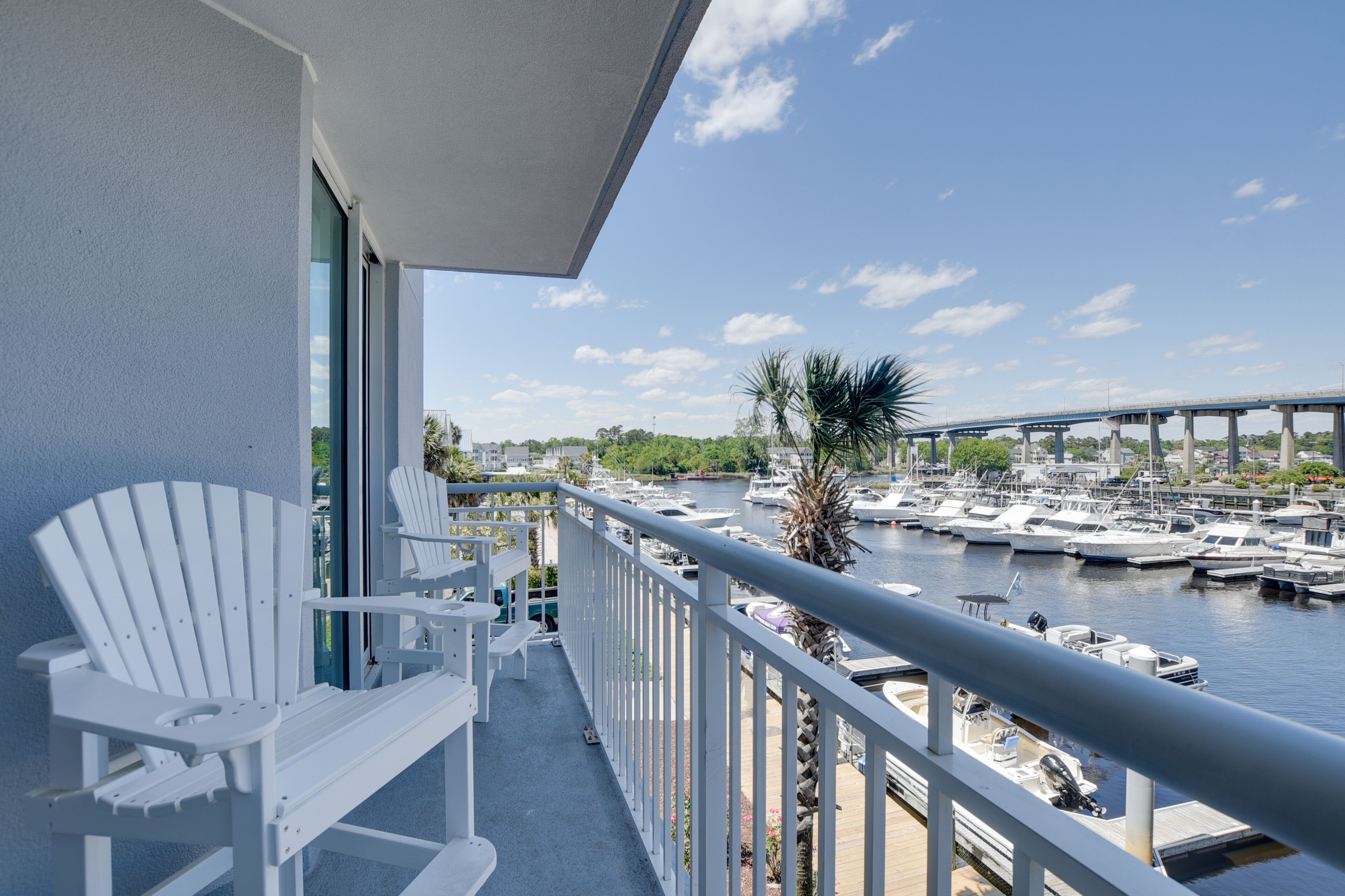 Un balcon avec des fauteuils Adirondack blancs, une vue sur une marina avec des bateaux, et un pont en arrière-plan.