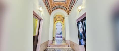 A hallway with a checkered floor, decorative ceiling, and wall sconces.