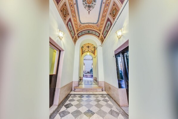 A hallway with a checkered floor, decorative ceiling, and wall sconces.