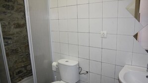 A bathroom with white tiled walls, a white toilet, a white sink, and a stone wall partially visible. - Encantaria SLU (Broto)