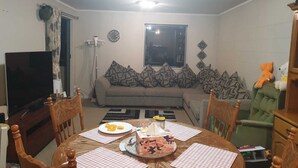 Ein Wohnzimmer mit einer Couch, einem Esstisch mit Platzsets, einem Fernseher und einer Uhr an der Wand.