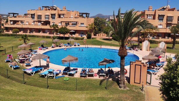 Un complejo turístico con una piscina central rodeada de tumbonas y sombrillas, y edificios al fondo.