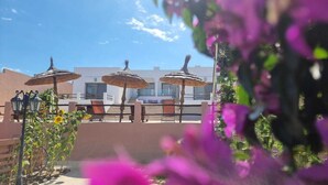 Un patio con mobiliario exterior y sombrillas, un edificio al fondo y algunas plantas en primer plano.