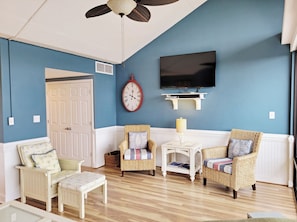 A room with a blue wall, a white door, a clock, a television, wicker chairs, a small table, and a bench.