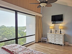 A bedroom with a large window, a ceiling fan, a television, a dresser, and a chair.