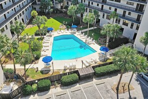Un espace piscine agrémenté de chaises longues et de parasols, entouré de palmiers et bordé d’un bâtiment doté de balcons.