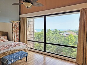 Une chambre avec une grande fenêtre, un lit, un banc et un ventilateur de plafond.