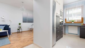 A modern kitchen with a refrigerator, oven, and microwave. There is a dining area with a blue sofa and a small table. A chandelier hangs from the ceiling, and a window provides natural light.