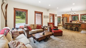 A spacious living room with a large sofa, a coffee table, and a red ottoman. There is a kitchen area with a bar and stools.