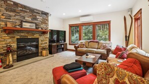 A cozy living room with a stone fireplace, a leather sofa, and a large window offering a view of trees.