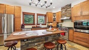 A modern kitchen with wooden cabinets, a large island with stools, a stone accent wall, and stainless steel appliances.