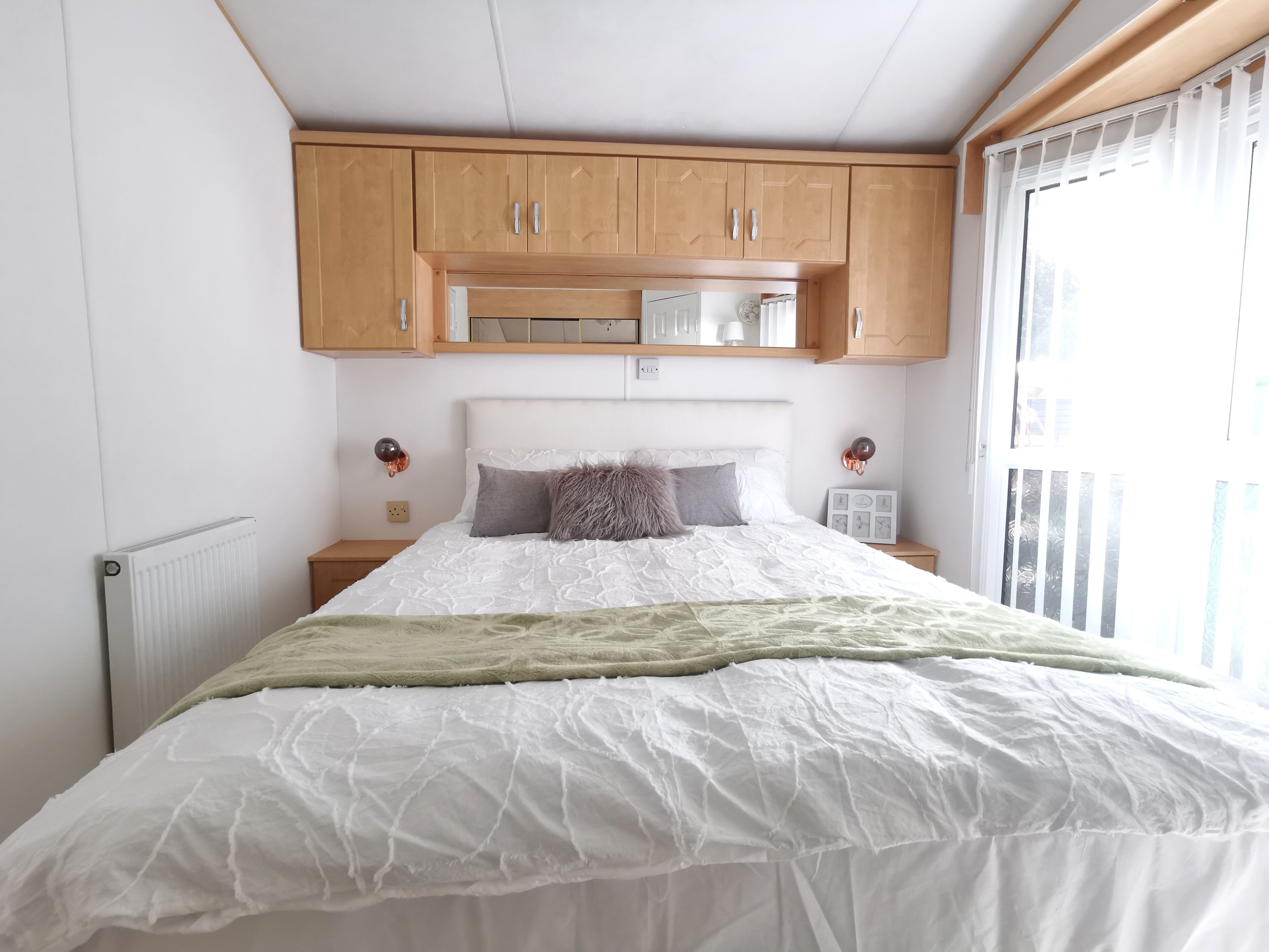 A bedroom with a bed, wooden cabinets, a radiator, a nightstand, and a window with blinds.