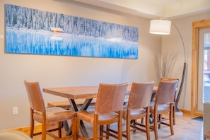 A dining area with a wooden table and chairs, a large painting on the wall, and a lamp. - Luxe Condo with Mountain Views, Balcony & BBQ (Canmore)