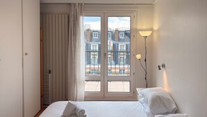Quarto de hotel com cama, janela com vista para edifícios, luminária de chão e radiador.