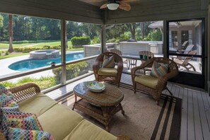 A covered outdoor area with wicker furniture, a glass table, and a view of a swimming pool and greenery.