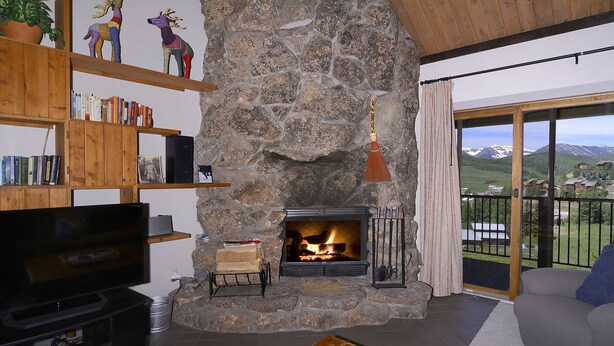A cozy living room with a stone fireplace, wooden shelves with books, a flat-screen TV, and a view of mountains through large glass doors. - Home Away From Home! Ski-in/Ski-out, Dog Friendly Condo w/ Fireplace & Views (Mt. Crested Butte)