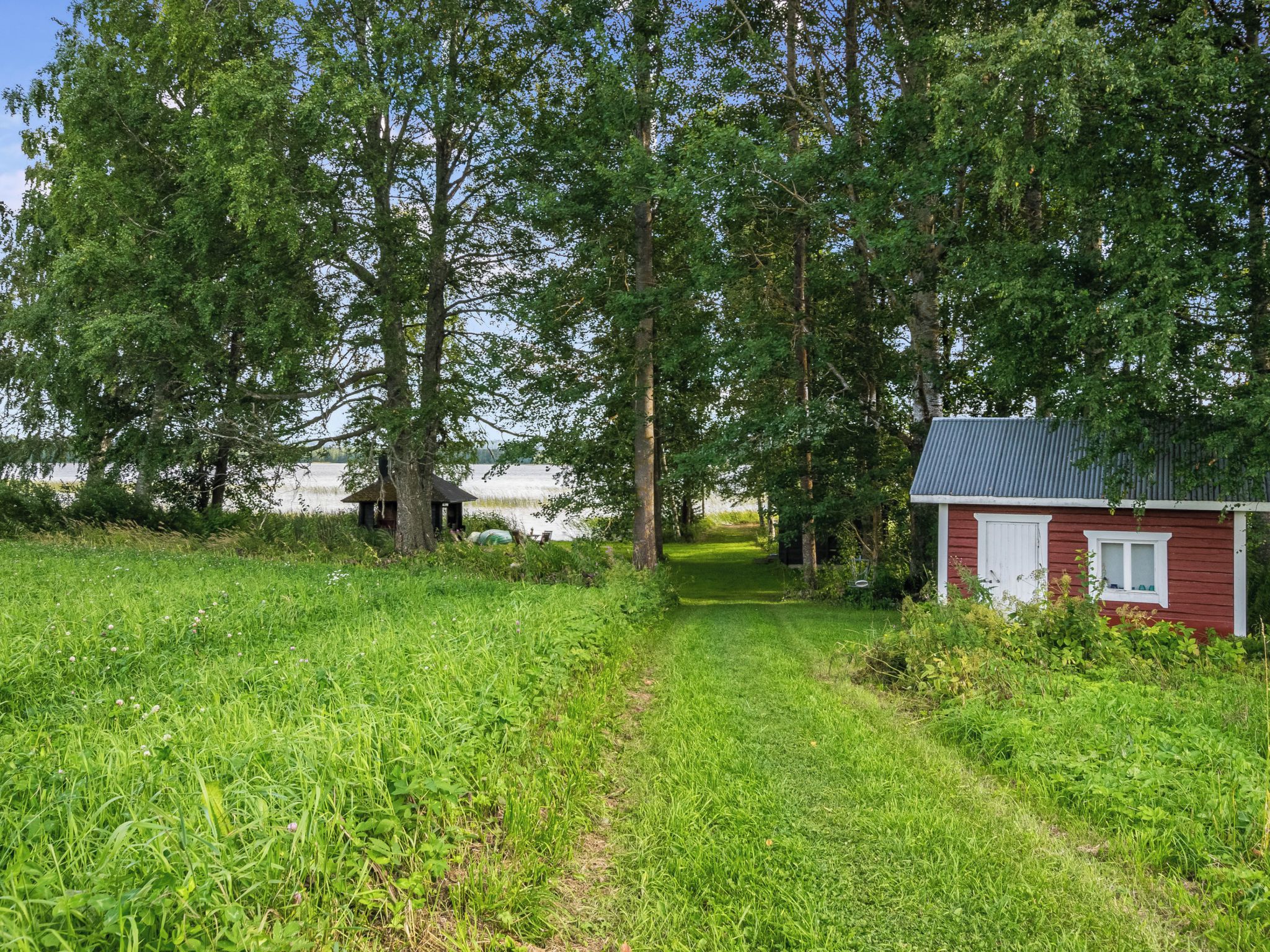 Un paysage rural avec une maison en bois rouge, un sentier qui traverse un champ d’herbe et de grands arbres.