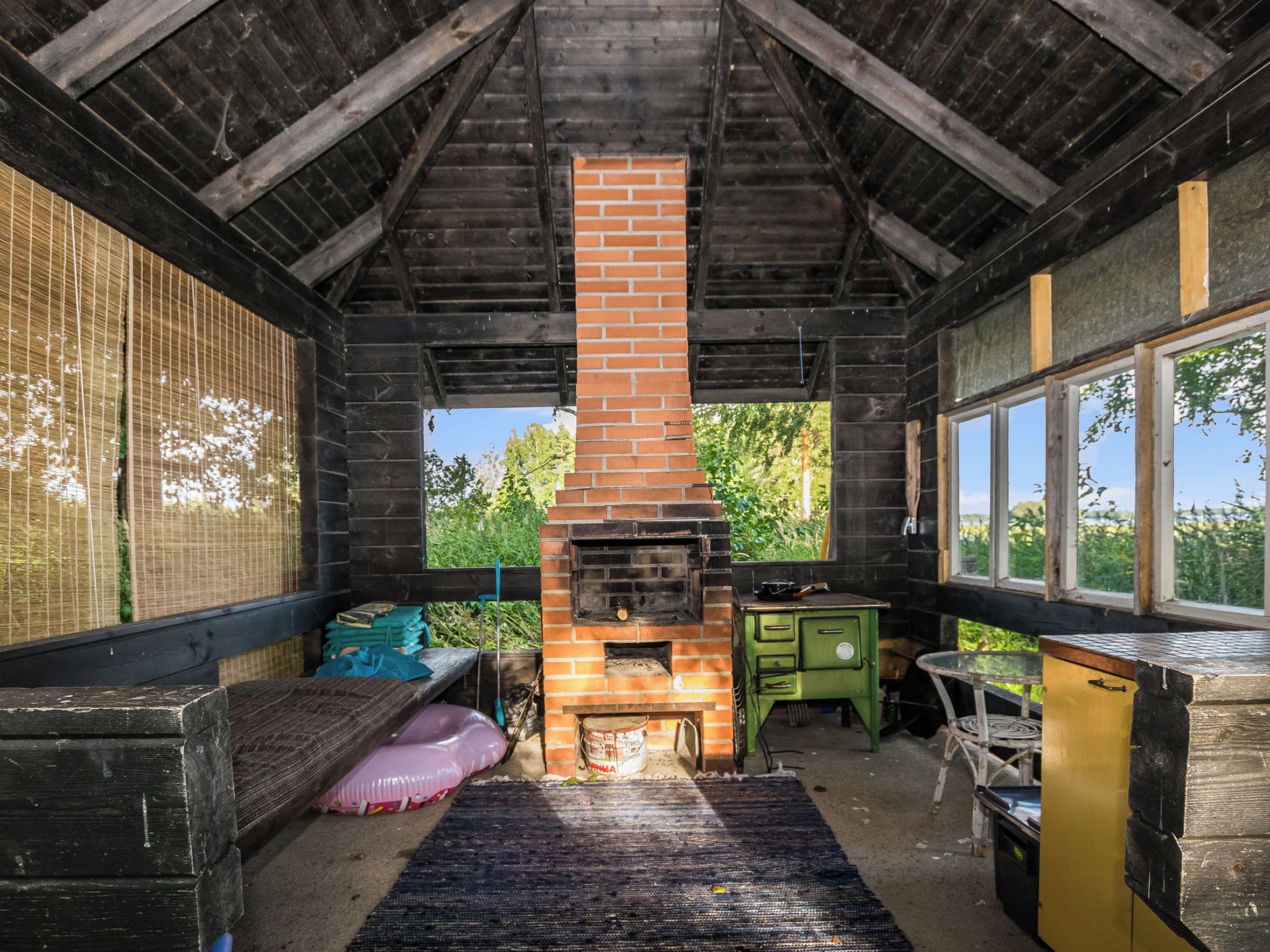 Une cabane en bois rustique avec une cheminée en briques, un poêle vert et une fenêtre donnant sur la verdure.