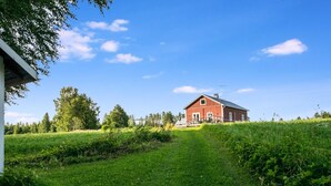 Une grange rouge avec une porte blanche et un petit porche, entourée de champs verdoyants et d’arbres sous un ciel bleu dégagé.
