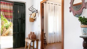 Quarto com piso de madeira, porta preta, parede branca com espelho, uma mesinha com dois vasos de barro e uma planta em vaso.