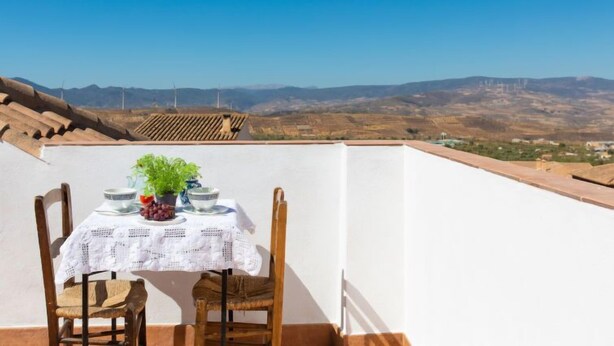 Mesa posta com toalha branca, uma tigela, uvas e uma planta em vaso num terraço com vista desobstruída para montanhas distantes e turbinas eólicas.