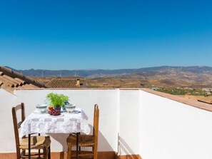 Una mesa cubierta con un mantel blanco, un cuenco, uvas y una planta en maceta en una terraza con vistas despejadas a montañas distantes y aerogeneradores.