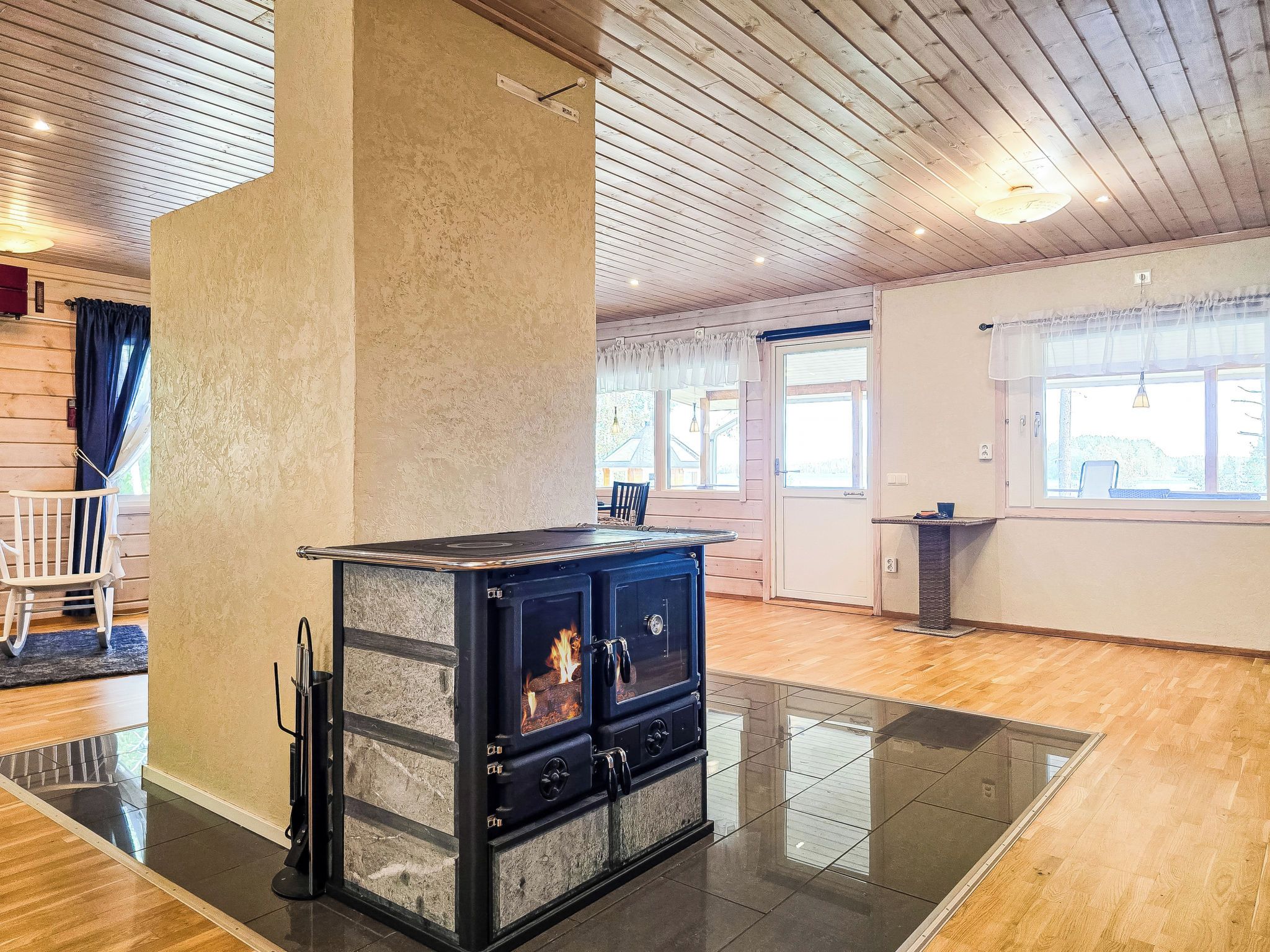 A room with a large, dark-colored wood-burning stove, a wooden ceiling, and a view through a window.