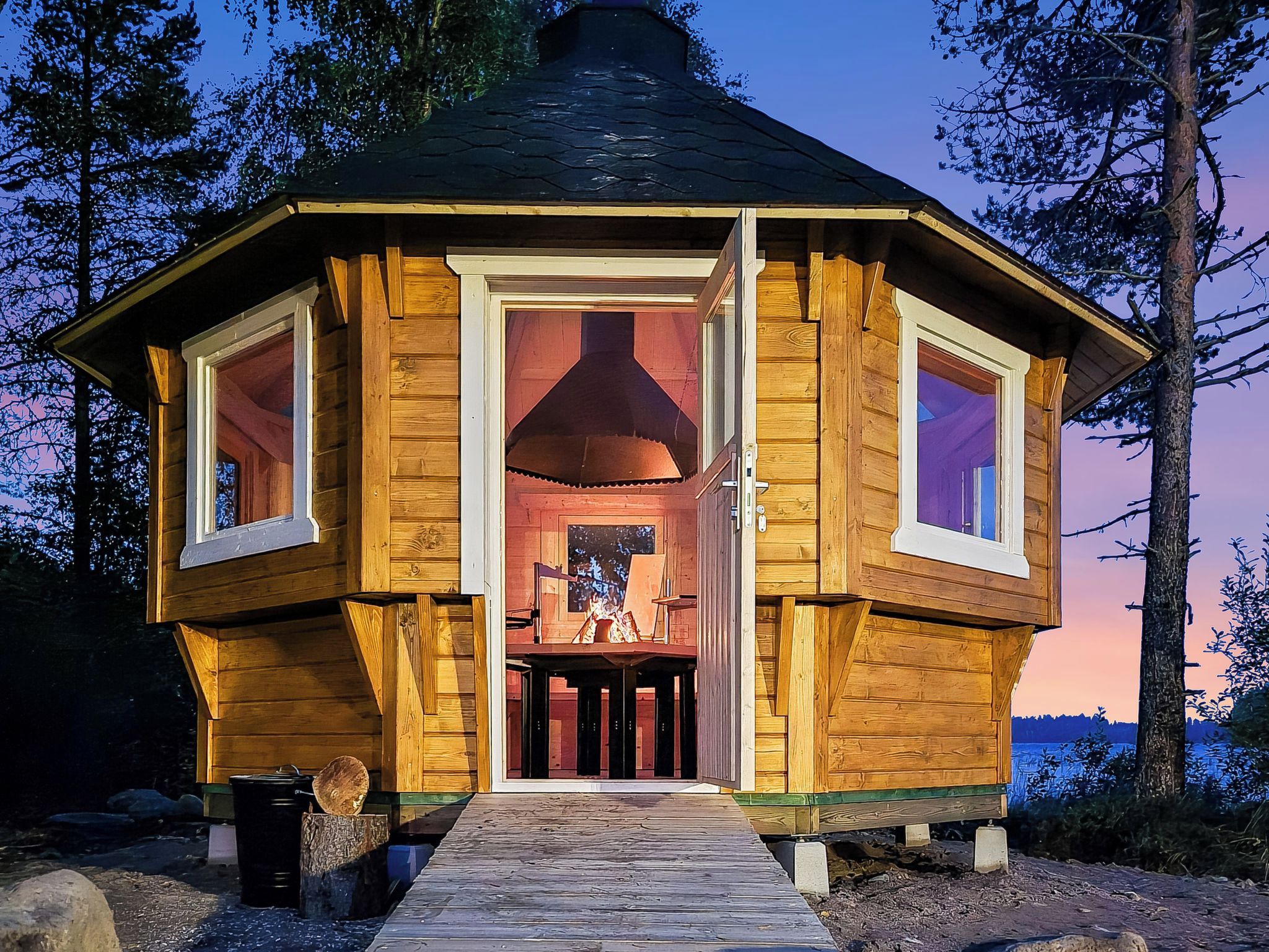 A wooden cabin with a central chimney, large windows, and a wooden deck leading to the entrance.