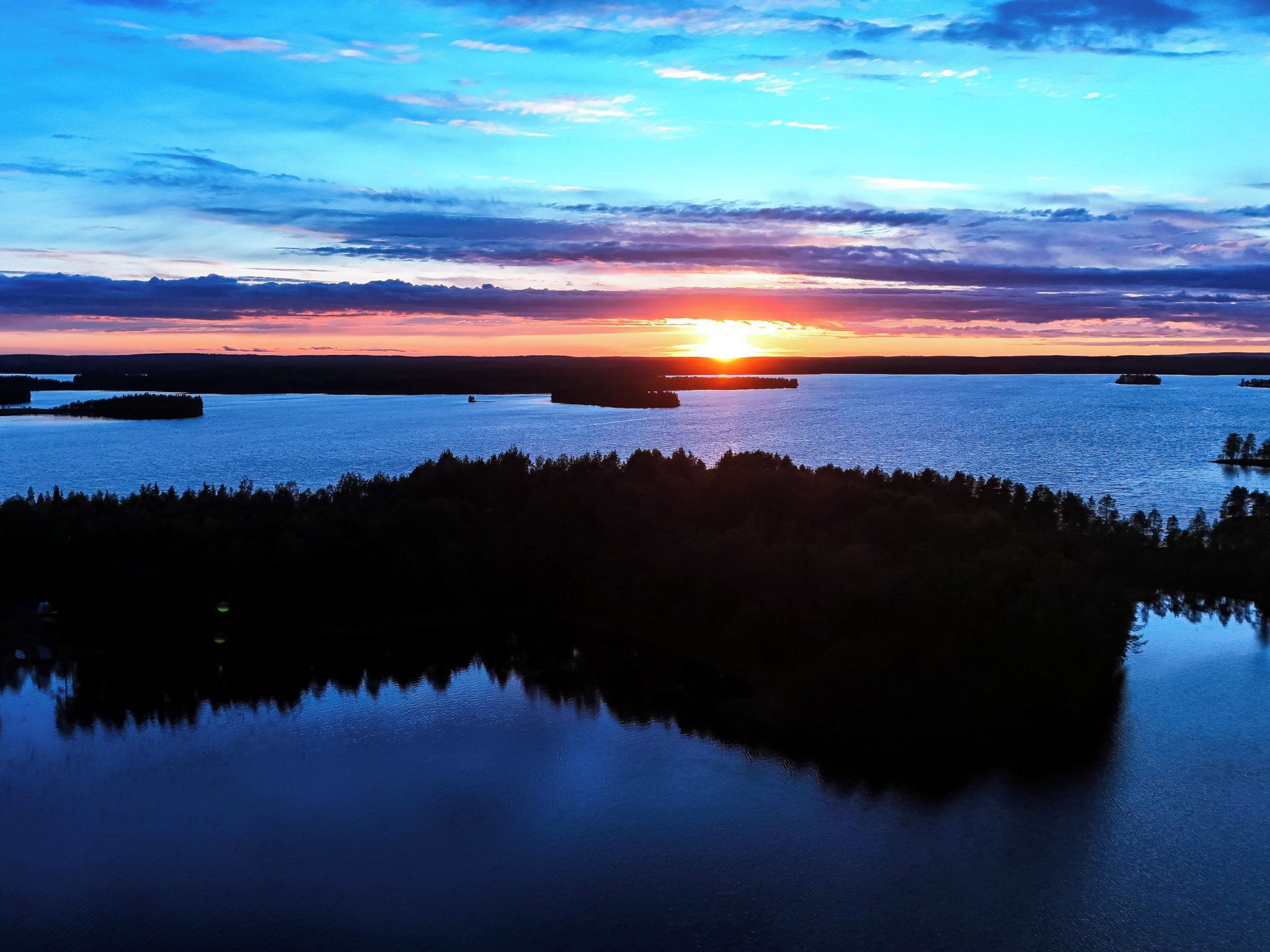 A calm lake at sunset with a forested shoreline and a boat in the distance.