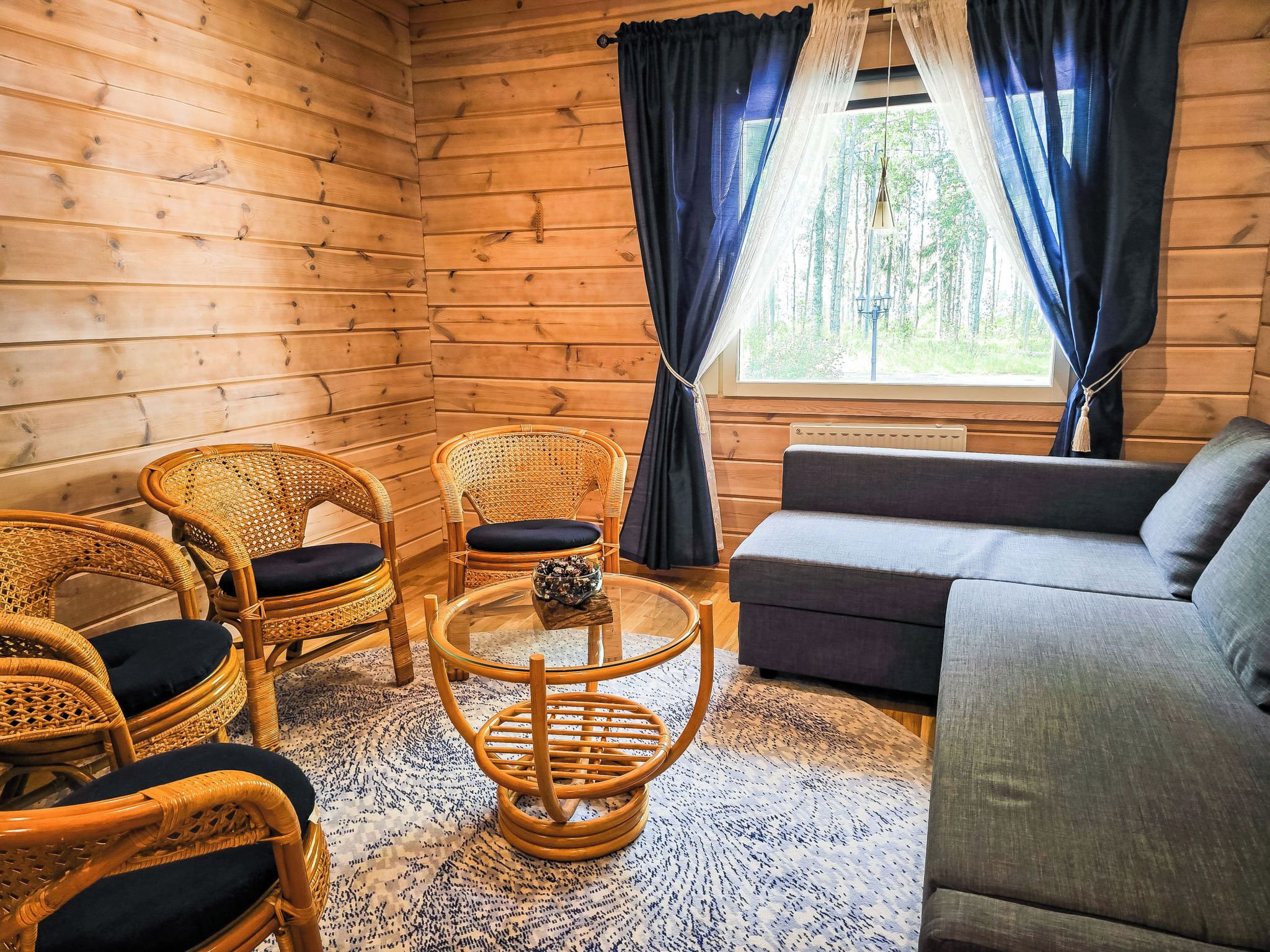 Interior acollidor d'una cabana amb parets de fusta, un sofà, dues cadires de vimetes, una taula de cafè de vidre i una vista del bosc a través d'una finestra amb cortines.