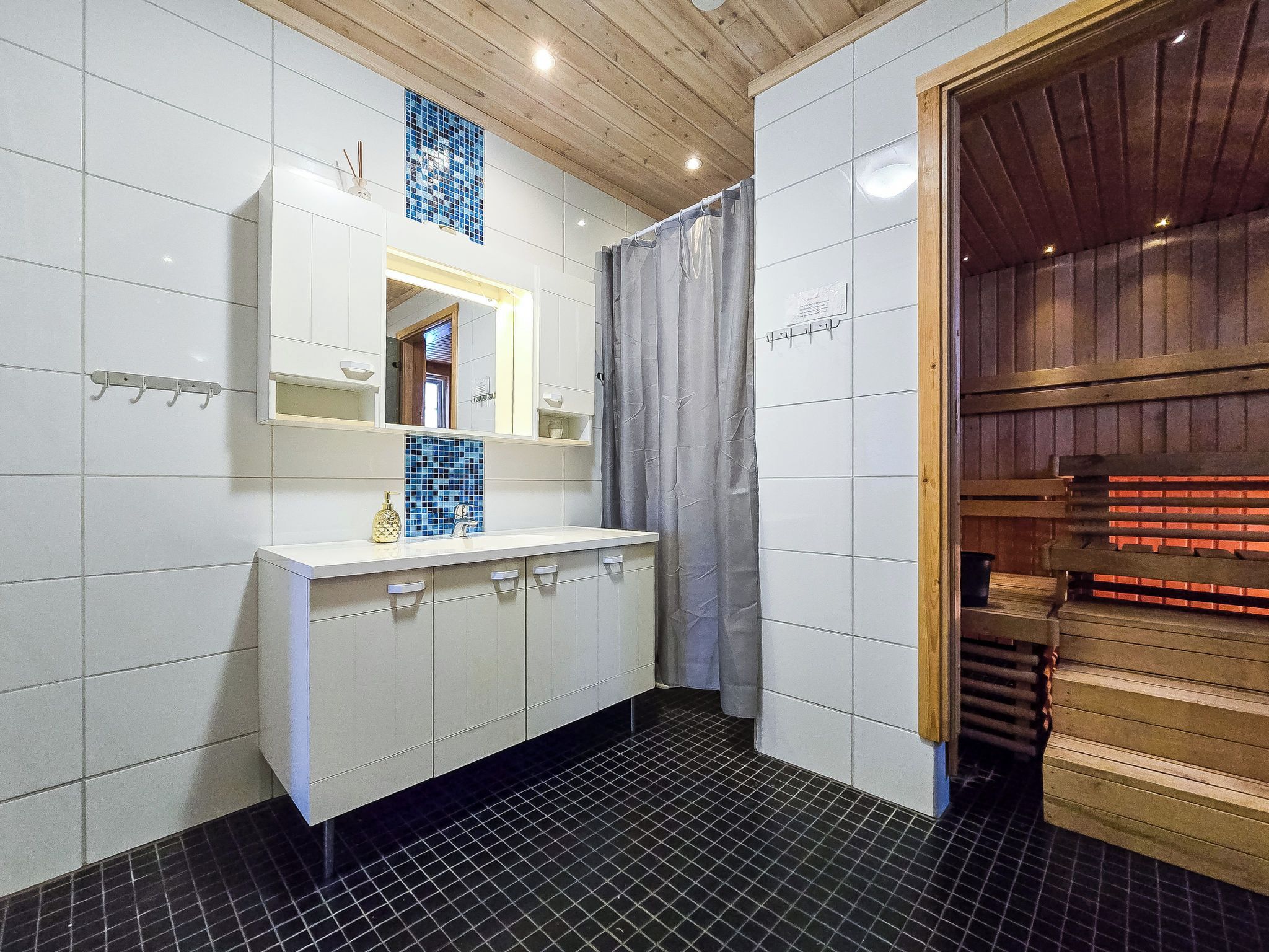 A bathroom with a white vanity, a mirror, a blue mosaic tile, a shower curtain, and a wooden sauna door.