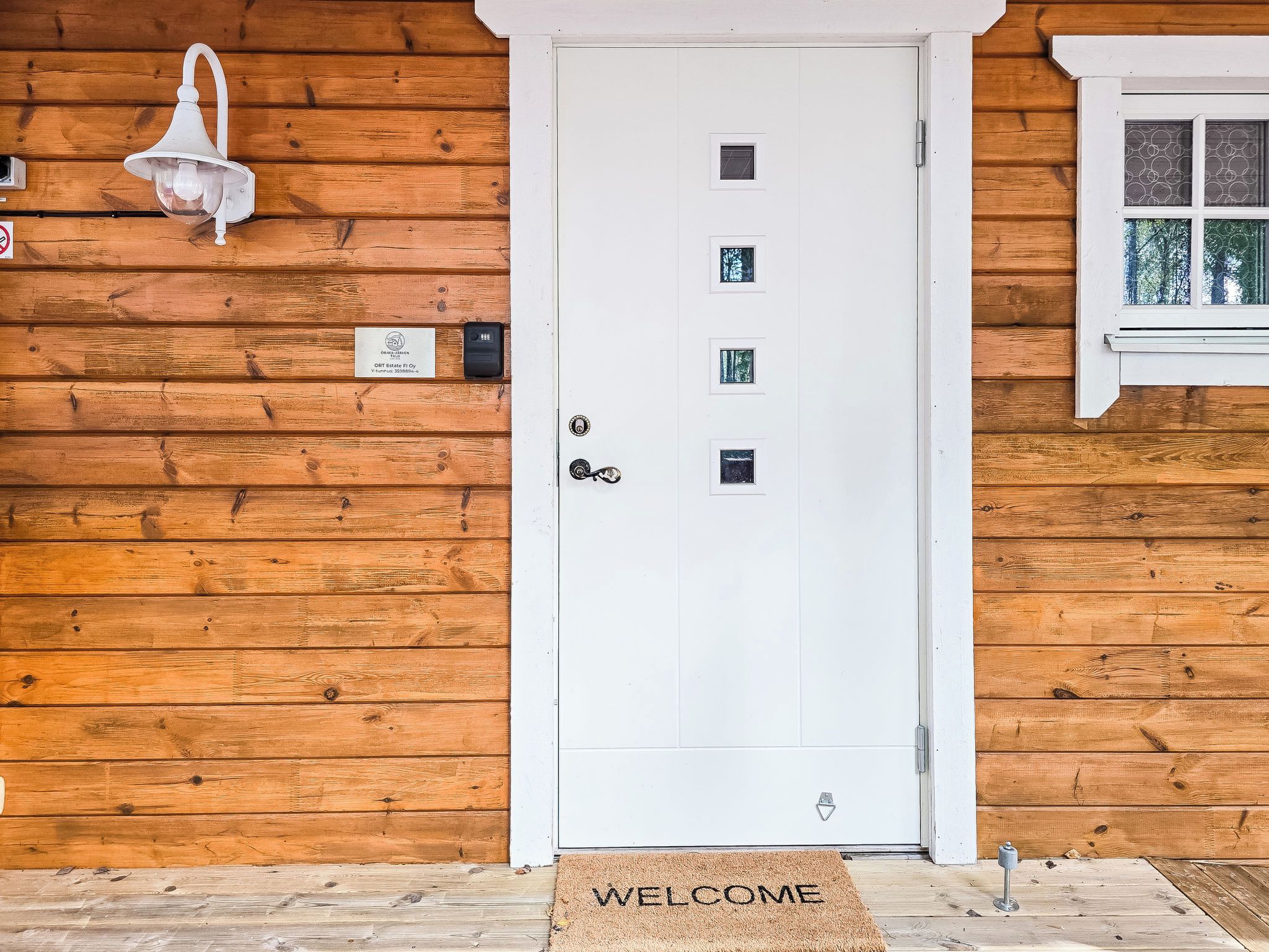 A white door with a doormat that reads 'WELCOME' and a small window with a view of trees.