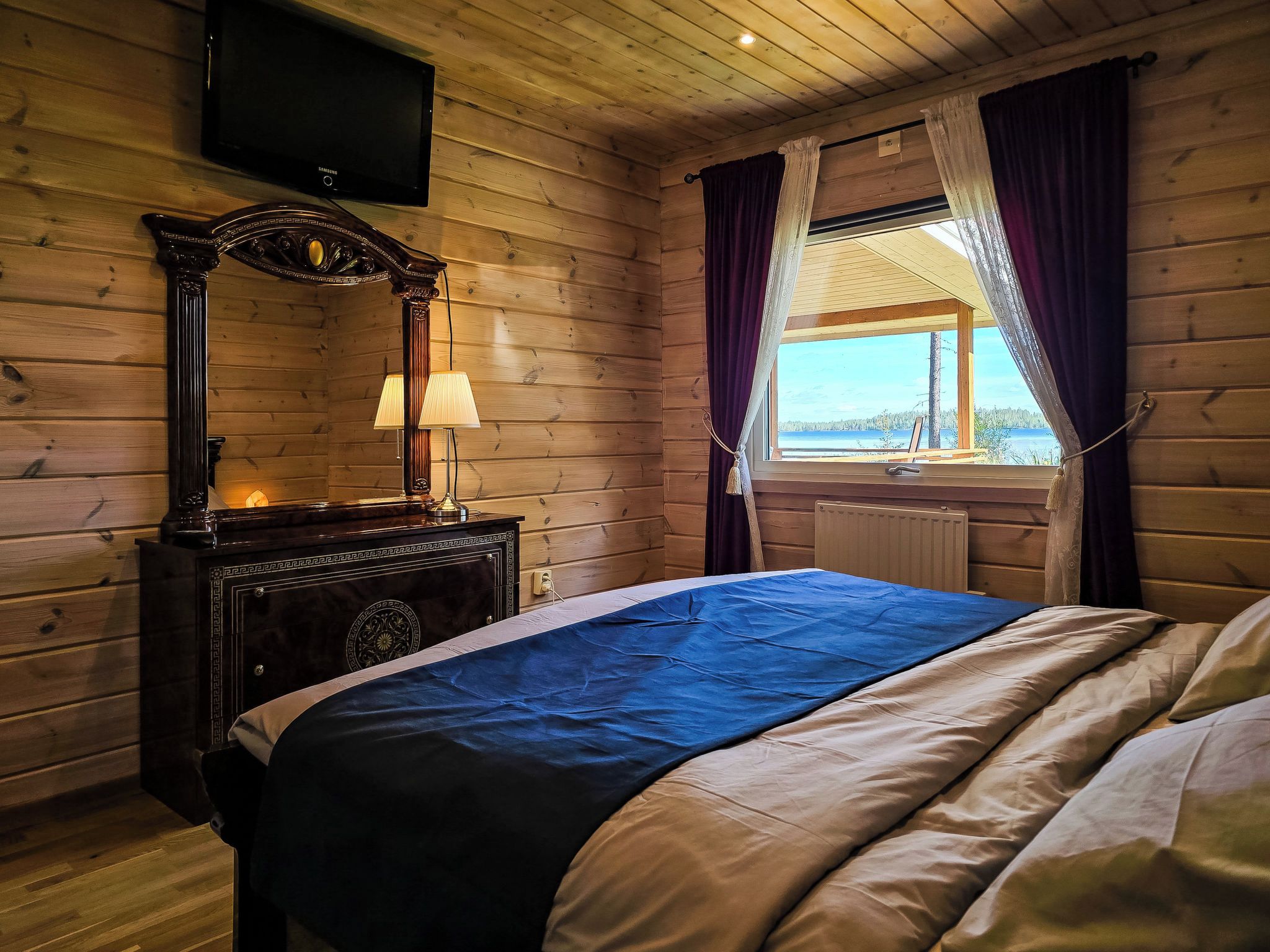 Una habitación de cabaña de madera con una cama, un aparador, un televisor y una ventana con cortinas.