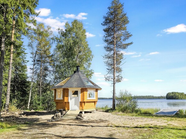 A small wooden cabin with a chimney, surrounded by trees and a lake.