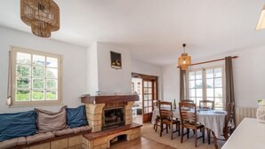 A cozy living room with a fireplace, a sofa, a dining table with chairs, and a large window. - Kergroix by Interhome (Quiberon)